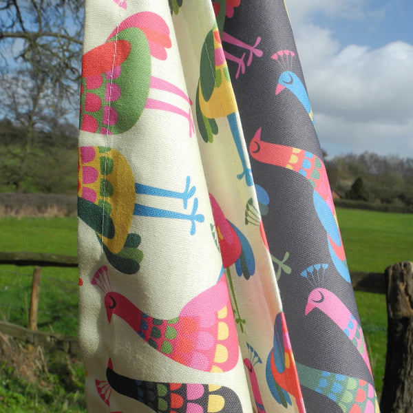 Two Haughty Peacocks tea towels shown hanging together on a washing line in the countryside