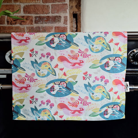 A Flowerbed tea towel by Rollerdog, with a colourful design comprising sighthounds, a squirrel and flowers, displayed over an oven door handle