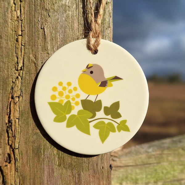 A ceramic hanging decoration illustrated with a goldcrest, shown hanging on a fencepost in the countryside