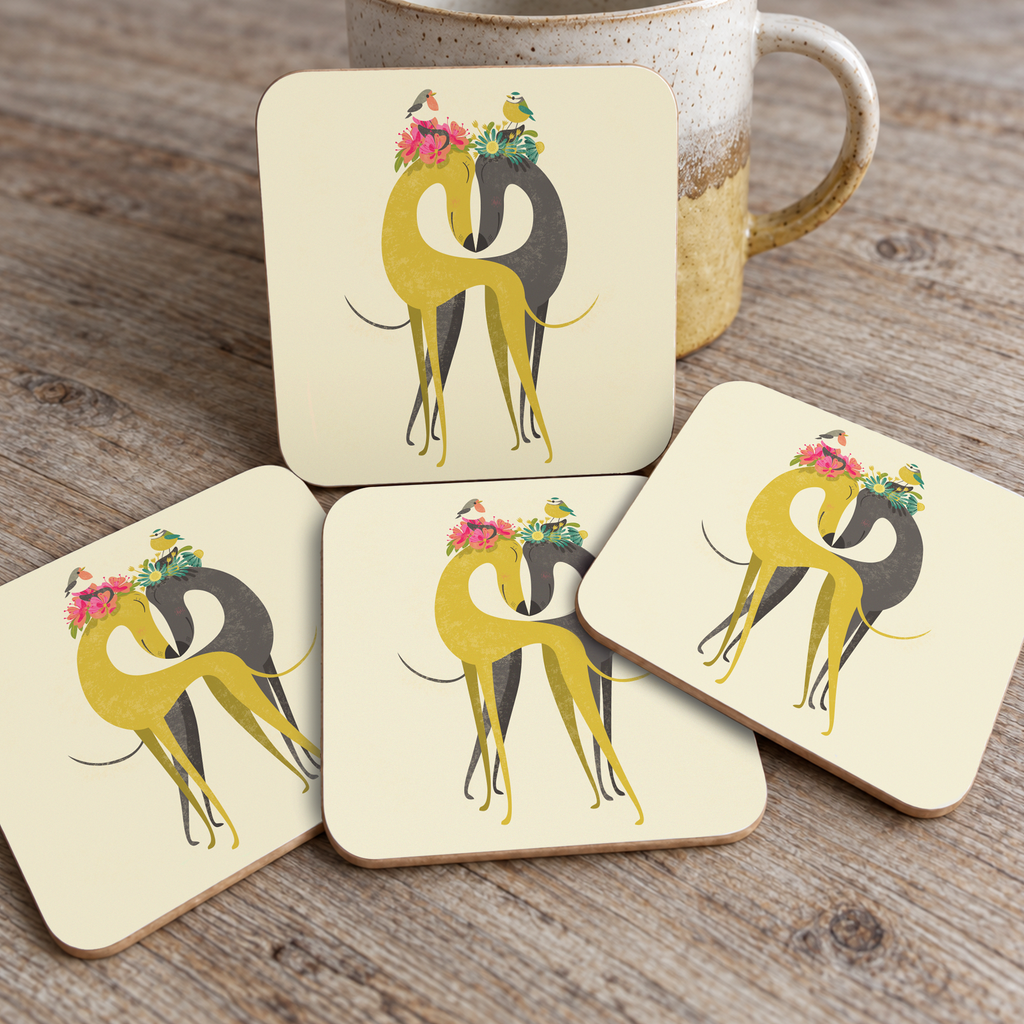Four square coaster tiles with rounded corners arranged on a wooden tabletop, with a ceramic mug partially visible behind them. Each coaster features a stylised yellow and black hound illustration with colourful flowers in their ears.