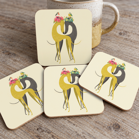 Four square coaster tiles with rounded corners arranged on a wooden tabletop, with a ceramic mug partially visible behind them. Each coaster features a stylised yellow and black hound illustration with colourful flowers in their ears.