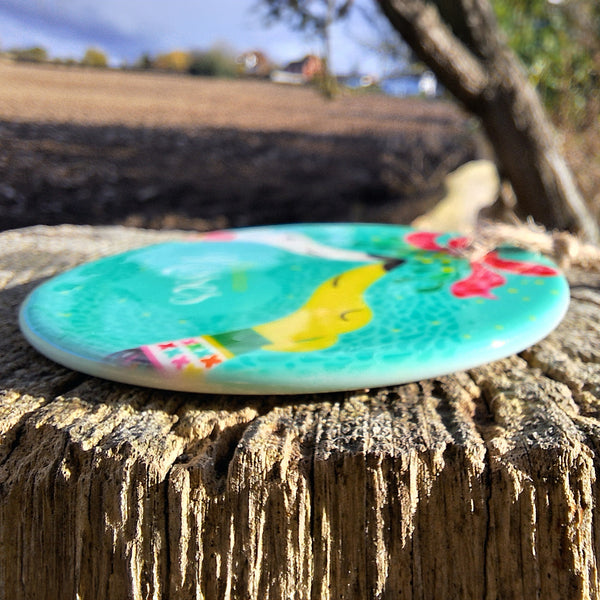 Side view of a flat ceramic decoration by Rollerdog, showing the thickness of the disk. The decoration is shown lying on a fencepost in the countryside.