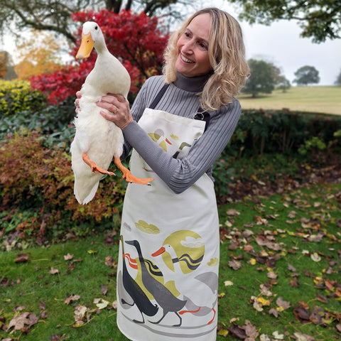 A  lady wearing a Rollerdog 'Derbyshire Ducks' pinny smiles at a white Indian runner duck, in a country garden setting