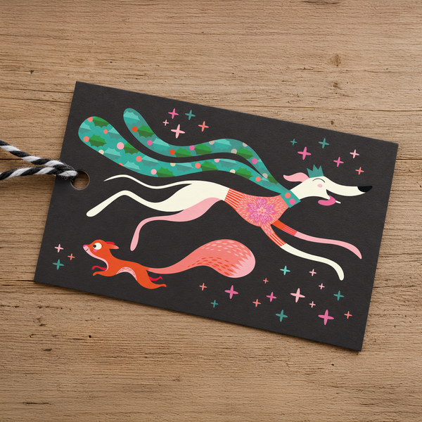 A black gift tag with a colourful illustration of a running sighthound, a squirrel, and small stars, attached to black-and-white striped twine, placed on a wooden surface.