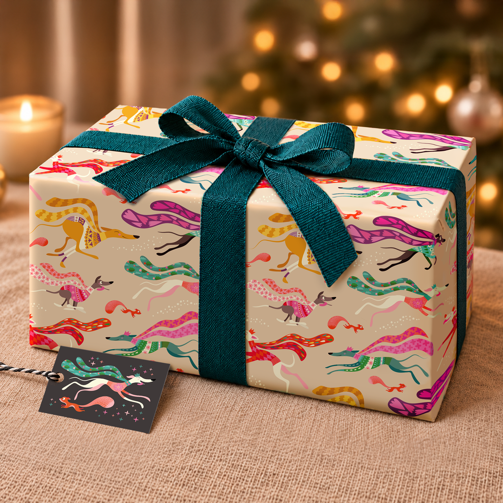A gift box wrapped in cream-coloured paper with a colourful sighthound and squirrel pattern, tied with a dark teal ribbon and bow, set on a flax surface with a blurred festive background.