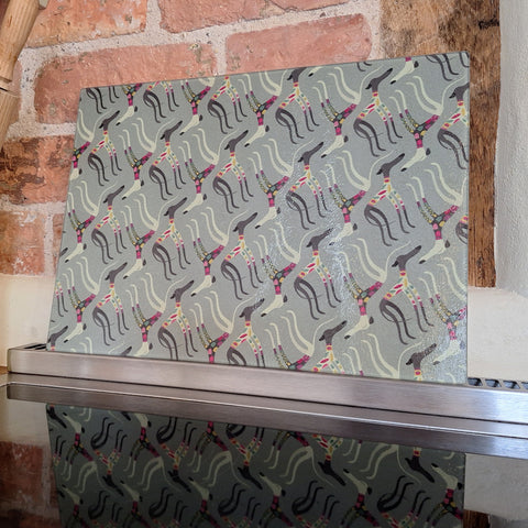 A glass chopping board with the Sleepy Sighthounds pattern, shown leaning against a brick wall on an oven hob