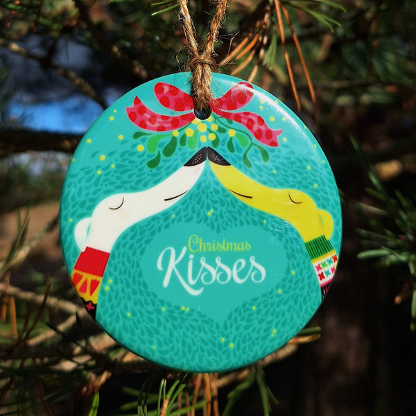 A flat ceramic disk decorated with artwork called Christmas Kisses, two hounds snout to snout under mistletoe