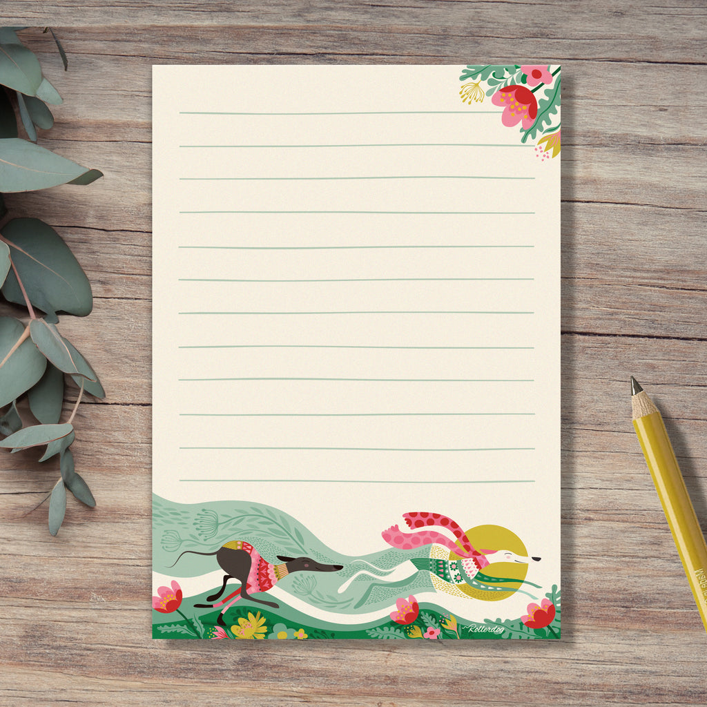 A Spring Zoomies notepad by Rollerdog, featuring an illustration of 2 hounds running fast in colourful jumpers,  through a bed of flowers. Eucalyptus leaves and a yellow pencil surround the notepad