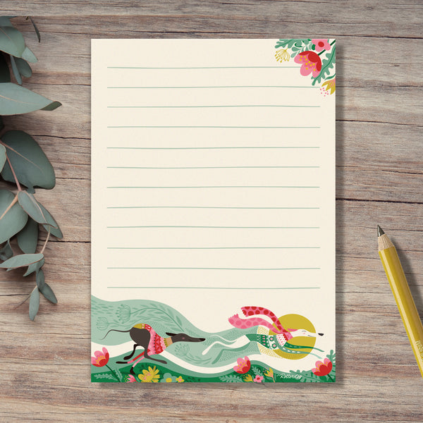 A Spring Zoomies notepad by Rollerdog, featuring an illustration of 2 hounds running fast in colourful jumpers,  through a bed of flowers. Eucalyptus leaves and a yellow pencil surround the notepad