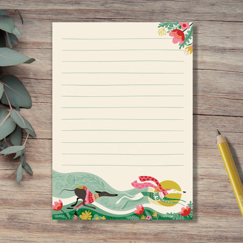 A Spring Zoomies notepad by Rollerdog, featuring an illustration of 2 hounds running fast in colourful jumpers,  through a bed of flowers. Eucalyptus leaves and a yellow pencil surround the notepad