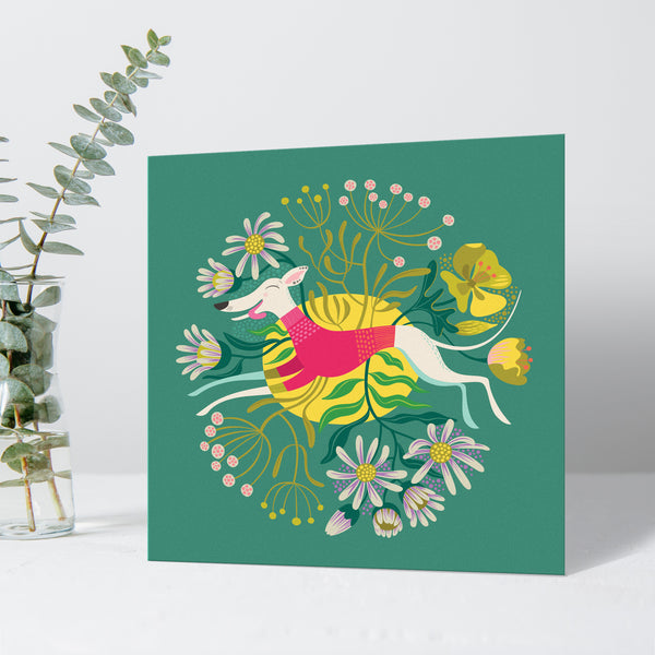 A  greeting card by Rollerdog featuring Snowy the whippet, a white hound leaping through seaside flora on a teal background