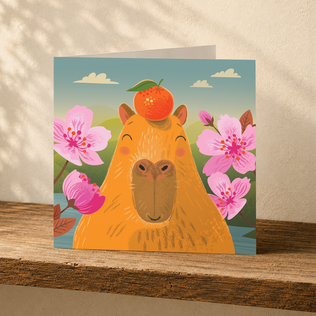 A square greeting card featuring a whimsical illustration of a capybara with an orange on its head, surrounded by pink flowers under a blue sky, displayed on a wooden surface against a textured beige wall.
