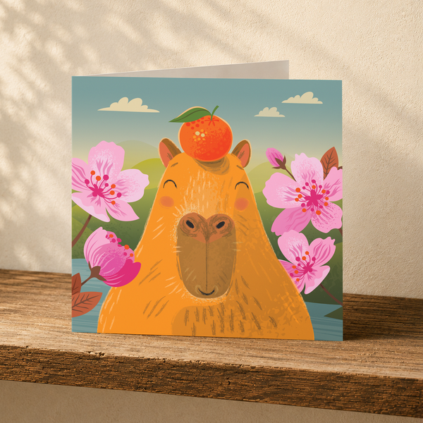 A square greeting card featuring a whimsical illustration of a capybara with an orange on its head, surrounded by pink flowers under a blue sky, displayed on a wooden surface against a textured beige wall.