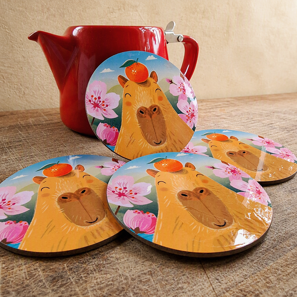A set of 4 coasters featuring a capybara illustration, on a wooden surface, leaning against a red teapot