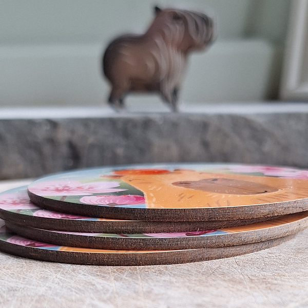 A close up view of a set of 4 capybara coasters on a wooden surface, with a cute toy capybara model in the background