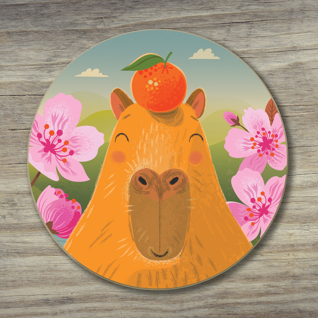 A round coaster featuring a whimsical illustration of a capybara with an orange fruit on her head, surrounded by pink cherry blossom flowers, placed on a grey wood-textured surface.