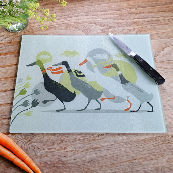 A Rollerdog Derbyshire Ducks glass chopping board, photographed from above on an oak worktop, with a paring knife, carrots and fresh flowers also in shot