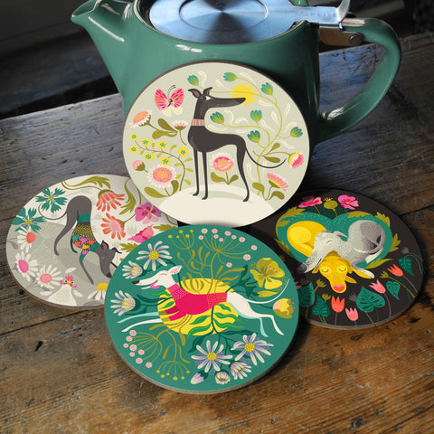 A set of four hardback coasters featuring greyhounds and whippets on colourful backgrounds, pictured against a teapot on a wooden table