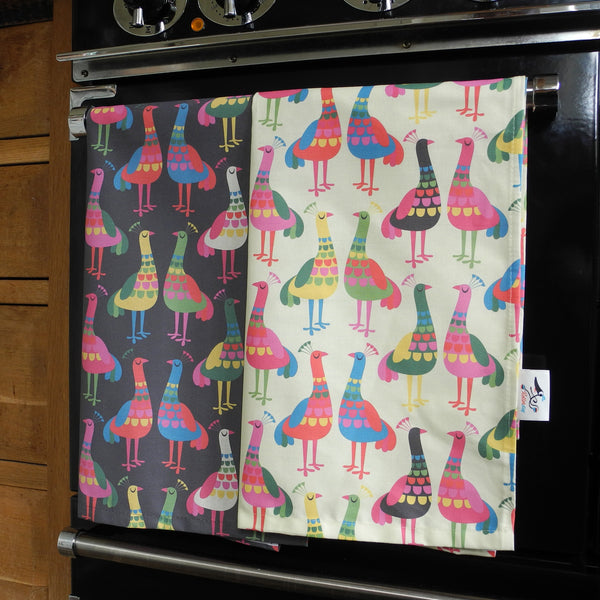 Two Haughty peacocks tea towels, showing the design on both the dark and light backgrounds. The tea towels are shown hanging on an oven door